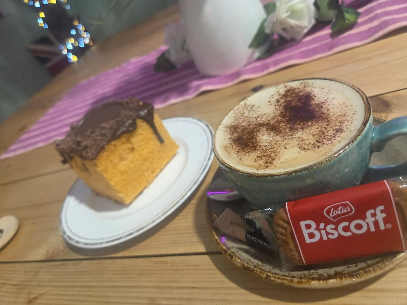 A cappuccino with a Biscoff biscuit next to a slice of cake